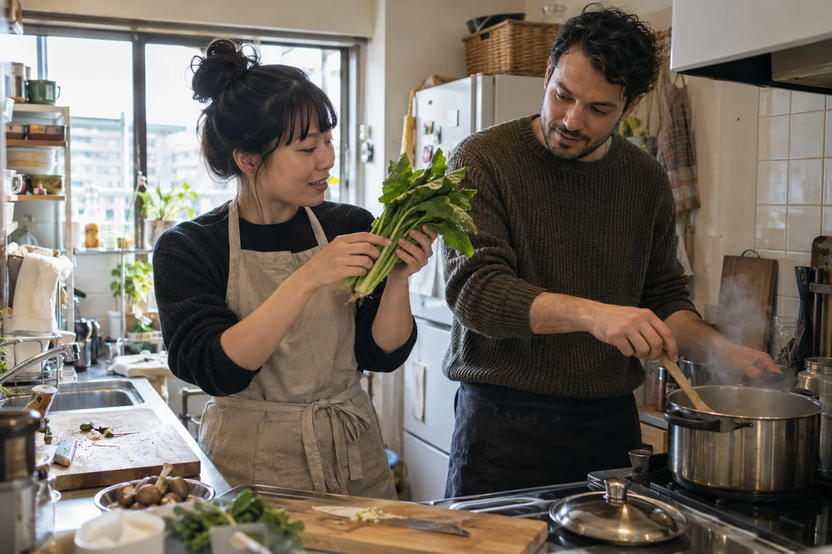 부드러운 햇살 아래 작은 주방에서 함께 요리하는 일본인 현지인과 외국인 거주자, 한 사람은 이곳의 잎채소 한 묶음을 들어 보이고 냄비에서는 김이 피어오름